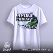 Футболка коли клює, біла, підвішена на вішалці, фон світло-сірий, принт з рибою та текстом "ДУША СПІВАЄ КОЛИ КЛЮЄ" в зелених та чорних кольорах.