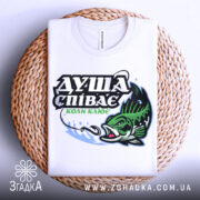 Футболка коли клює, біла, складена на плетеній основі, фон світло-сірий, принт з рибою та текстом "ДУША СПІВАЄ КОЛИ КЛЮЄ" в зелених та чорних кольорах.