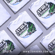 Футболка коли клює, біла, зображена на манекені, фон світло-сірий, принт з рибою та текстом "ДУША СПІВАЄ КОЛИ КЛЮЄ" в зелених та чорних кольорах.