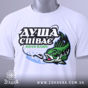 Футболка коли клює, біла, складена на поверхні, фон світло-сірий, принт з рибою та текстом "ДУША СПІВАЄ КОЛИ КЛЮЄ" в зелених та чорних кольорах, з етикеткою на шнурку.