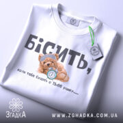 Футболка коли тебе будять, біла футболка, складена на столі, ведмедик з годинником на принті, ракурс зверху, фон світлий, текст чорний і сірий, на футболці є етикетка з зеленим шнурком.