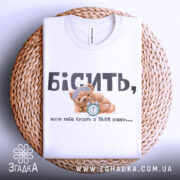 Футболка коли тебе будять, біла футболка, складена на плетеному кошику, ведмедик з годинником на принті, ракурс зверху, фон натуральний, текст чорний і сірий.