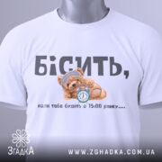 Футболка коли тебе будять, біла футболка, складена на столі, ведмедик з годинником на принті, ракурс зверху, фон світлий, текст чорний і сірий, на футболці є етикетка з зеленим шнурком.