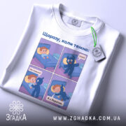 Футболка коли темно, біла, зображена зверху, на ній є етикетка, фон світлий, принт аналогічний першому фото, з чотирма коміксами, що зображують діалог між хлопчиком і синім персонажем.