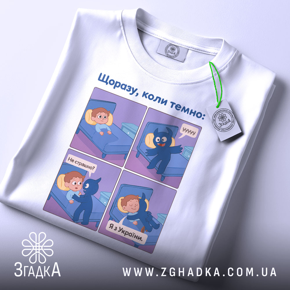 Футболка коли темно, біла, зображена зверху, на ній є етикетка, фон світлий, принт аналогічний першому фото, з чотирма коміксами, що зображують діалог між хлопчиком і синім персонажем.