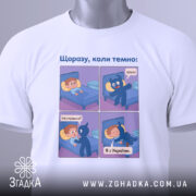 Футболка коли темно, біла, зображена зверху, на ній є етикетка, фон світлий, принт аналогічний першому фото, з чотирма коміксами, що зображують діалог між хлопчиком і синім персонажем.