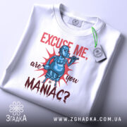 Футболка комічний маніяк, біла, складена на поверхні, фон світло-сірий, принт з персонажем у масці, що тримає трубку, текст червоним кольором, з етикеткою на рукаві.