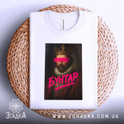 Футболка король бунтар, біла, зображена зверху, на фоні плетеного кошика, принт з портретом короля, з рожевими мазками на очах та написом 'БУНТАР', з видимою етикеткою.