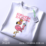 Футболка королева хламінго, біла, складена на плоскій поверхні, на фоні світло-сірого кольору, принт з рожевим фламінго в короні, що тримає коктейль, з зеленим ярликом.