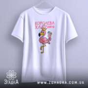 Футболка королева хламінго, біла, підвішена на вішалці, на фоні світло-сірого кольору, принт з рожевим фламінго в короні, що тримає коктейль, з чіткими деталями.