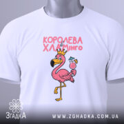 Футболка королева хламінго, біла, складена на плоскій поверхні, на фоні світло-сірого кольору, принт з рожевим фламінго в короні, що тримає коктейль, з зеленим ярликом.
