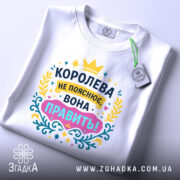Футболка королева білого кольору, зображена зверху на плоскій поверхні, фон світлий, принт яскравий з текстом "КОРОЛЕВА НЕ ПОЯСНЮЄ, ВОНА ПРАВИТЬ!" в кольорах жовтому, рожевому, блакитному та зеленому, з етикеткою на комірці.