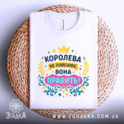 Футболка королева білого кольору, зображена зверху на плоскій поверхні, фон світлий, принт яскравий з текстом "КОРОЛЕВА НЕ ПОЯСНЮЄ, ВОНА ПРАВИТЬ!" в кольорах жовтому, рожевому, блакитному та зеленому, розташована на плетеній основі.