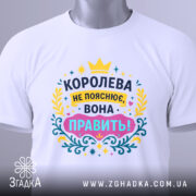 Футболка королева білого кольору, зображена зверху на плоскій поверхні, фон світлий, принт яскравий з текстом "КОРОЛЕВА НЕ ПОЯСНЮЄ, ВОНА ПРАВИТЬ!" в кольорах жовтому, рожевому, блакитному та зеленому, з етикеткою на комірці.