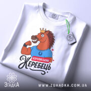 Футболка королівський жеребець, білого кольору, лежить на плоскій поверхні, фон світло-сірий, принт з конем у короні, що демонструє м'язи, з написом 'Королівський жеребець', на футболці є етикетка з зеленим шнурком.