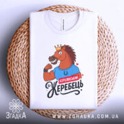 Футболка королівський жеребець, білого кольору, лежить на плетеній кошику, фон світло-сірий, принт з конем у короні, що демонструє м'язи, з написом 'Королівський жеребець', видно етикетку на комірці.