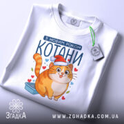 Футболка котани з новим роком, біла, зображена зверху, фон світло-сірий, принт з котом у червоній шапці, на футболці є етикетка з зеленим шнурком.