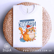 Футболка котани з новим роком, біла, зображена на плетеній кошику, фон білий, принт з котом у червоній шапці, футболка складена.