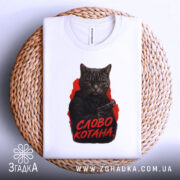 Футболка котяча мафія, біла футболка складена на плетеній кошику, фон світло-сірий, принт з котом у чорному капюшоні з пістолетом, текст 'СЛОВО КОТАНА' червоним кольором.