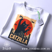 Футболка котяча монстр білого кольору, зображена зверху, на фоні світло-сірого, принт з чорним котом, що піднімає лапу на фоні червоного сонця, з зеленим ярликом.