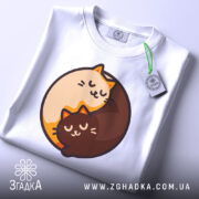 Футболка котячий інь-янь, біла, зображена зверху, на столі, фон світло-сірий, принт з двома котами, один кремового кольору, інший коричневий, обидва з закритими очима, на футболці є етикетка з зеленим шнурком.