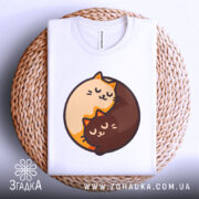 Футболка котячий інь-янь, біла, зображена зверху на плетеній кошику, фон світло-сірий, принт з двома котами, один кремового кольору, інший коричневий, обидва з закритими очима, виглядають спокійно.