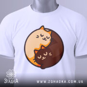 Футболка котячий інь-янь, біла, зображена зверху, на столі, фон світло-сірий, принт з двома котами, один кремового кольору, інший коричневий, обидва з закритими очима, на футболці є етикетка з зеленим шнурком.