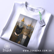 Футболка коти Київ, біла, складена на поверхні, фон світлий, принт з двома котами, на футболці є етикетка з зеленим шнурком.