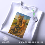 Футболка коти на велосипедi, біла, зображена зверху, на фоні світло-сірого градієнту, принт з двома котами на велосипеді, один сірий, інший рудий, обидва з усмішками, на фоні зеленого пейзажу, з етикеткою на комірці.