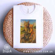 Футболка коти на велосипедi, біла, складена на плетеній кошику, на фоні світло-сірого градієнту, принт з двома котами на велосипеді, один сірий, інший рудий, обидва з усмішками, на фоні зеленого пейзажу.