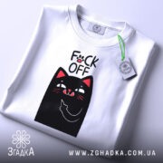 Футболка котик фак офф, складена футболка білого кольору, з принтом чорного кота, який показує середній палець, фон світло-сірий, на комірці є етикетка.