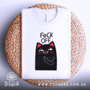 Футболка котик фак офф, складена футболка білого кольору, з принтом чорного кота, який показує середній палець, фон світло-сірий, футболка розташована на плетеній основі.