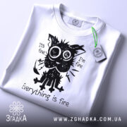 Футболка котик гумор, біла футболка на столі, ракурс зверху, фон світло-сірий, принт з котиком з великими очима, текст "Everything is fine", на футболці зелена етикетка.