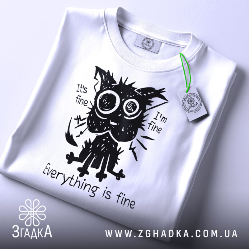 Футболка котик гумор, біла футболка на столі, ракурс зверху, фон світло-сірий, принт з котиком з великими очима, текст "Everything is fine", на футболці зелена етикетка.