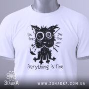Футболка котик гумор, біла футболка на столі, ракурс зверху, фон світло-сірий, принт з котиком з великими очима, текст "Everything is fine", на футболці зелена етикетка.