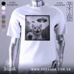 Футболка котик і НЛО, біла футболка на манекені, зображення кота з відкритим ротом і піднятою лапою, на фоні видно НЛО, принт в чорній рамці, фон світлий.