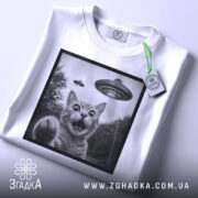 Футболка котик і НЛО, складена біла футболка з принтом кота, на фоні світло-сірий, на футболці є етикетка з зеленим шнурком, принт в чорній рамці.