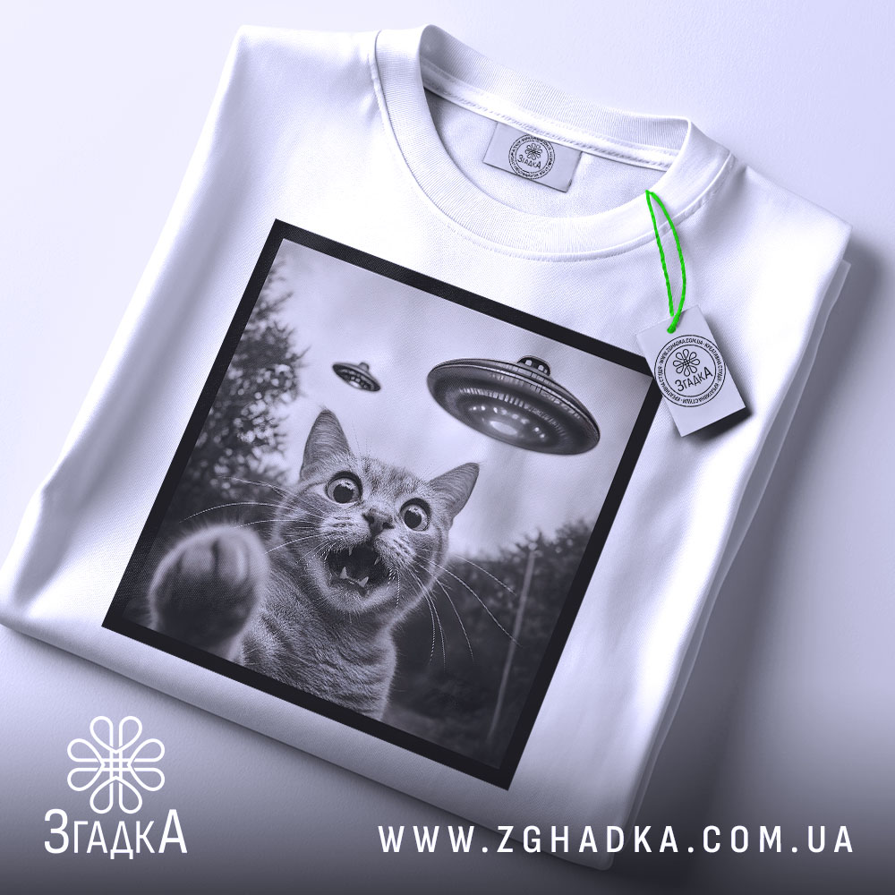 Футболка котик і НЛО, складена біла футболка з принтом кота, на фоні світло-сірий, на футболці є етикетка з зеленим шнурком, принт в чорній рамці.