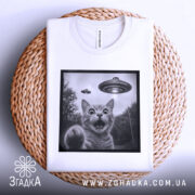 Футболка котик і НЛО, складена біла футболка на плетеній основі, зображення кота з відкритим ротом і піднятою лапою, на фоні видно НЛО, принт в чорній рамці.