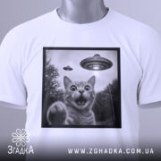 Футболка котик і НЛО, складена біла футболка з принтом кота, на фоні світло-сірий, на футболці є етикетка з зеленим шнурком, принт в чорній рамці.