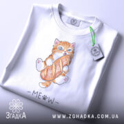 Футболка котик з батоном, біла, складена на поверхні, фон світлий, принт з котиком, що тримає батон, з написом 'MEOW' під ним, на футболці є етикетка з зеленим шнурком.