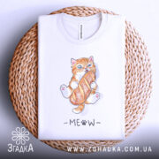Футболка котик з батоном, біла, складена на плетеній кошику, фон світлий, принт з котиком, що тримає батон, з написом 'MEOW' під ним.