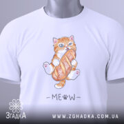Футболка котик з батоном, біла, складена на поверхні, фон світлий, принт з котиком, що тримає батон, з написом 'MEOW' під ним, на футболці є етикетка з зеленим шнурком.