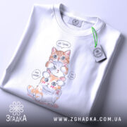 Футболка котики кумедні, біла, складена на столі, фон світло-сірий, принт з трьома котиками, один оранжевий, один білий, один з оранжевими плямами, з написами 'No Speak', 'No Hear', 'No See', на ярлику зелена стрічка.