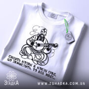 Футболка козак грає, біла футболка, зображена зверху, принт козака з кобзою, на футболці є етикетка з зеленим шнурком, фон світлий, деталі принта яскраві.