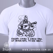 Футболка козак грає, біла футболка, зображена зверху, принт козака з кобзою, на футболці є етикетка з зеленим шнурком, фон світлий, деталі принта яскраві.