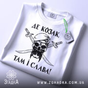 Футболка козак і слава, біла футболка, складена на поверхні, зображена з верхнього ракурсу, фон світло-сірий, принт чорного кольору з черепом у беретці та двома мечами, текст українською мовою, на футболці є етикетка з зеленим шнурком.