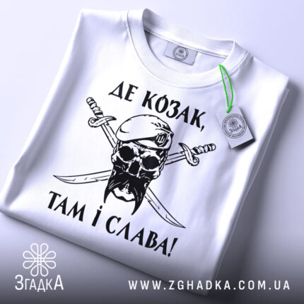 Футболка козак і слава, біла футболка, складена на поверхні, зображена з верхнього ракурсу, фон світло-сірий, принт чорного кольору з черепом у беретці та двома мечами, текст українською мовою, на футболці є етикетка з зеленим шнурком.