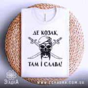 Футболка козак і слава, біла футболка, складена на плетеній кошику, зображена з верхнього ракурсу, фон світло-сірий, принт чорного кольору з черепом у беретці та двома мечами, текст українською мовою, видима етикетка на комірці.