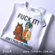 Футболка козак не жени. Біла футболка, складена на столі, з принтом, що зображує скелета в українському вбранні на коні. На фоні світло-сірий градієнт. Текст на футболці: 'FUCK IT! DON'T PUSH THE HORSES', з етикеткою на комірці.